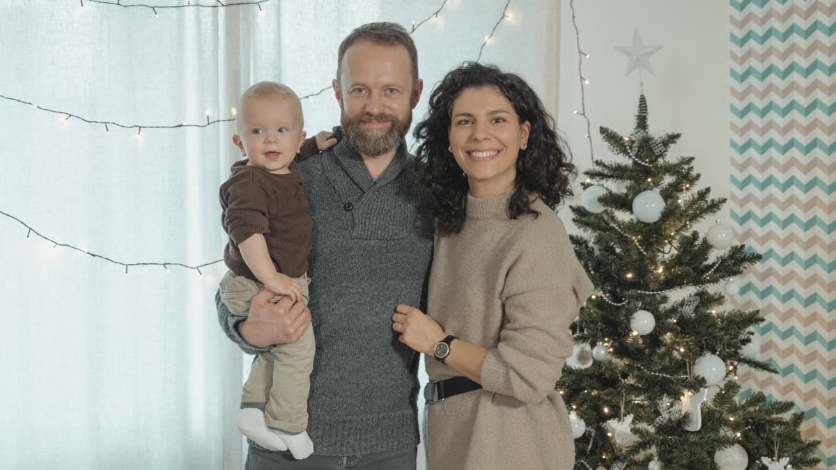 A joyful family with a baby poses next to a decorated Christmas tree indoors.